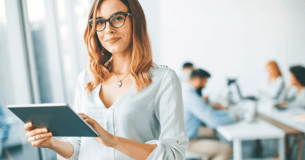 Una mujer autónoma profesional sonríe mientras gestiona las facturas y calcula qué es el IRPF de su actividad en una tablet, con su equipo de trabajo de fondo en la oficina.