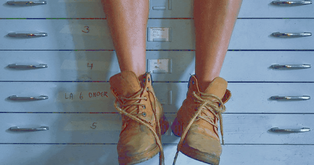 Primer plano de unas piernas con botas de trabajo colgadas frente a unos archivadores metálicos de oficina numerados.