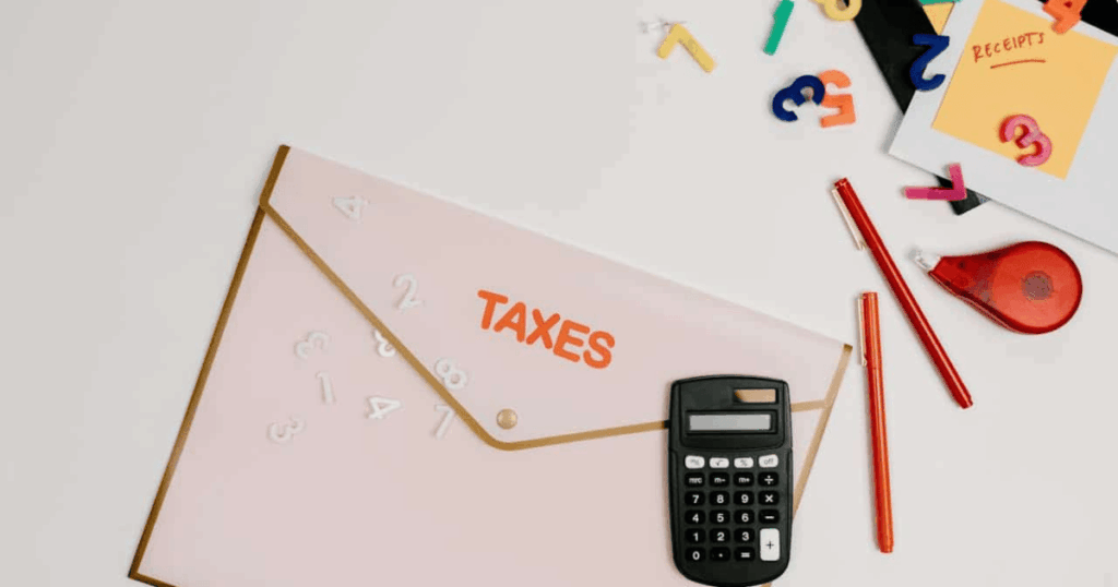 Vista cenital de un escritorio con una carpeta rosa con la palabra TAXES (impuestos), una calculadora, un bolígrafo rojo y números sueltos, ilustrando cómo un autónomo gestiona sus gastos para deducir el IVA de una factura simplificada.