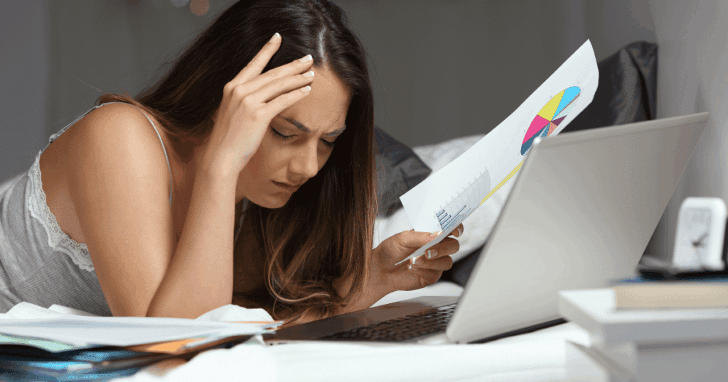 Una mujer autónoma preocupada y con dolor de cabeza revisa documentos fiscales y gráficos frente a su portátil por la noche.