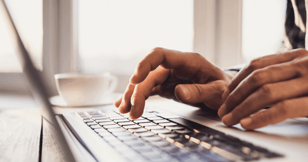 Primer plano de unas manos escribiendo en el teclado de un ordenador portátil plateado sobre una mesa de madera, con una taza de café borrosa al fondo, ilustrando la acción de instalar el certificado digital en otro ordenador.