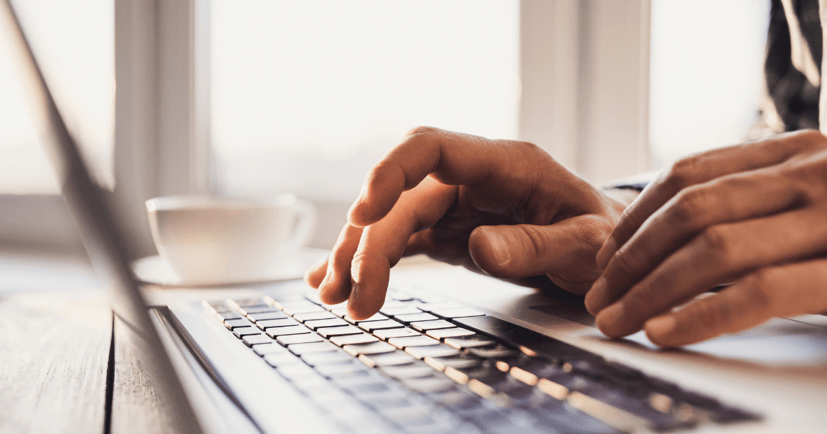 Primer plano de unas manos escribiendo en el teclado de un ordenador portátil plateado sobre una mesa de madera, con una taza de café borrosa al fondo, ilustrando la acción de instalar el certificado digital en otro ordenador.